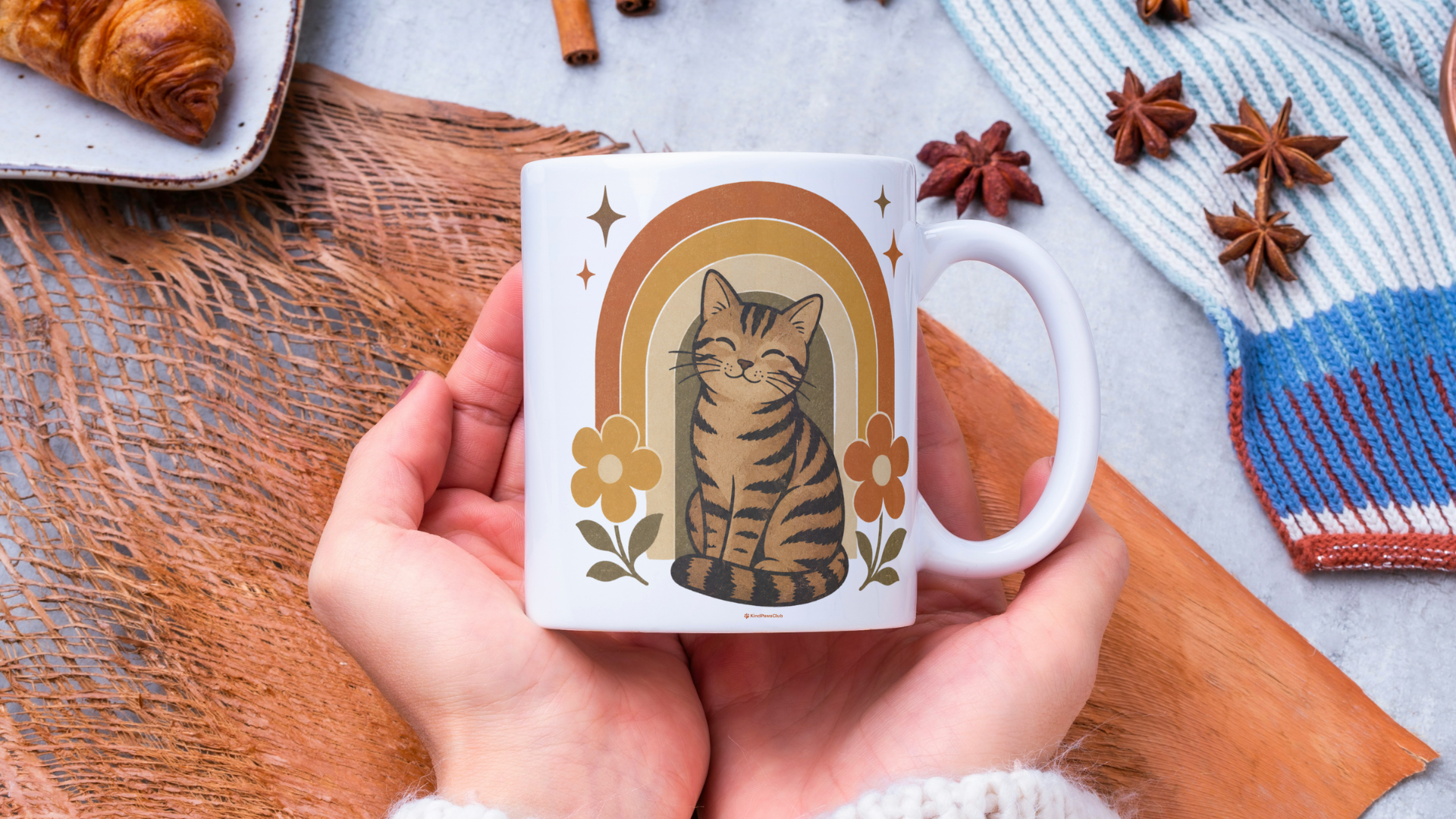 Person holding a mug with a cat illustration on a wooden surface with cinnamon sticks and a croissant.